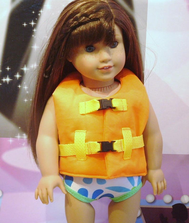 May include: A doll wearing an orange life vest with yellow straps and blue and white patterned swim trunks. The doll has long brown hair and blue eyes.