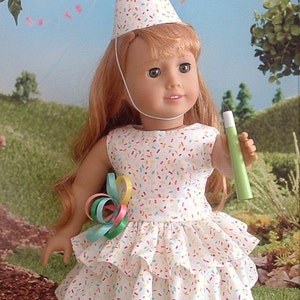 Op de afbeelding: Een pop die een witte feestmuts draagt met kleurrijke sprinkels en een witte jurk met kleurrijke sprinkels. De pop houdt een groen feestcadeau vast.