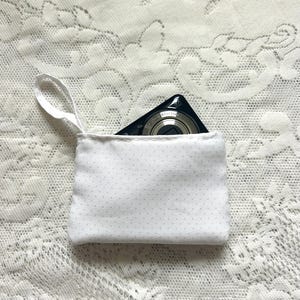 May include: A small white fabric pouch with a subtle grey polka dot pattern and a matching white wrist strap. A black digital camera is partially tucked inside the pouch, with the lens and top edge of the camera body visible above the opening.