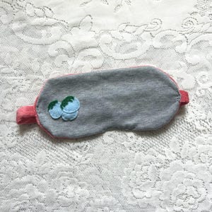 May include: A grey sleep mask with a pink trim and a small appliqué of two light blue berries with green stems on the left side. The mask features a matching pink elastic strap on each side for securing it around the head during use.