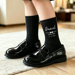 May include: Black ribbed crew socks featuring the white printed text Daniel, a small bow tie icon, the words PAGE BOY, and the date 21.12.2026. The socks are worn with black patent leather loafers and black shorts, reaching mid-calf in height.