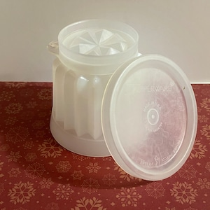 May include: A clear, vintage Tupperware container with a lid. The container is cylindrical with vertical ridges and a textured lid. The lid is partially open, revealing the interior. The text "Tupperware" and "Made in USA" are visible on the lid.