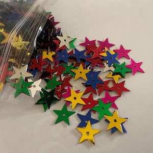 May include: A clear plastic bag filled with colorful star-shaped sequins. The sequins are in various colors, including red, yellow, green, blue, and silver. These decorative embellishments are scattered on a white surface.