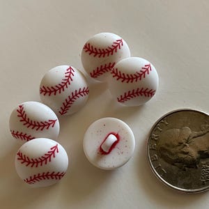 May include: A collection of small, baseball-themed decorative buttons. The buttons are white with red stitching, resembling miniature baseballs. A quarter is included for size comparison.