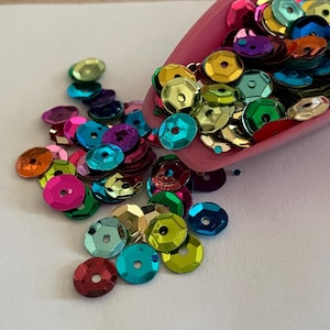 May include: A pink scoop overflowing with colorful, iridescent sequins. The sequins are in various colors, including pink, blue, green, gold, and purple. They are scattered on a white surface, creating a vibrant and decorative display.