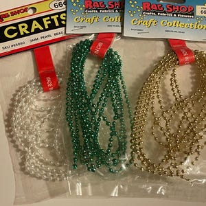 May include: Three packages of 3mm pearl bead strands from Rag Shop Craft Collection. Each clear plastic bag contains a continuous string of beads in white, green, or gold. The header cards feature the text Rag Shop Crafts and Craft Collection.