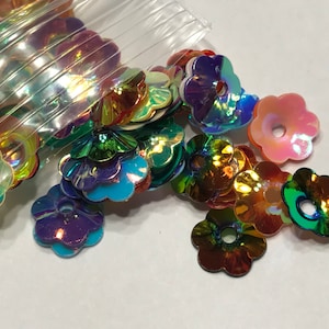 May include: Assortment of iridescent flower-shaped sequins in various colors, including pink, green, blue, and orange. The sequins are scattered on a white surface.
