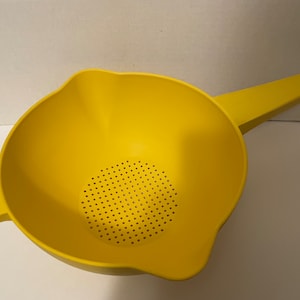 May include: A bright yellow plastic colander featuring a long handle with a circular hole at the end, a smaller side handle with a circular hole, and a perforated base for draining liquids. The bowl has a wide rim and a pouring spout on the side.