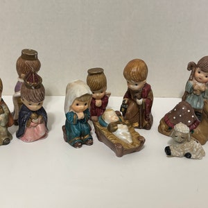 May include: A set of ten ceramic nativity figurines featuring stylized figures including a baby in a manger, figures in robes of blue, red, and brown, a camel, and a sheep. Each piece has painted facial features and muted color tones.