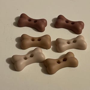 May include: Six bone-shaped buttons in various shades of brown and beige. Each button features two holes in the center for attachment. The buttons have a smooth, matte finish and are arranged in two rows of three on a plain surface.