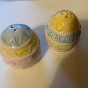 May include: Two ceramic egg-shaped salt and pepper shakers. The shakers are painted in pastel colors, including yellow, blue, and pink. Each shaker has a different design, with floral and patterned details. The tops have holes for dispensing.