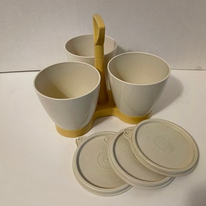 May include: A vintage Tupperware serving caddy in cream and yellow. The caddy has three cone-shaped containers with lids, and a handle for easy carrying. The lids and containers are cream-colored, and the caddy is yellow.