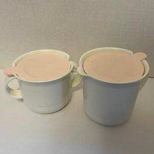 May include: Two white mugs with handles, each topped with a light pink lid. The lids have a small tab for easy removal. The mugs are set against a plain white background.