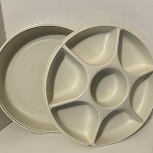 May include: Off-white, round serving tray with a lid. The tray has a divided section with six compartments and a central well. The lid is a simple, shallow dish. The tray is made of a durable plastic material.