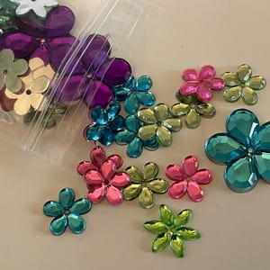 May include: A collection of colorful flower-shaped embellishments in various shades of pink, green, blue, purple, and gold. Some are loose, while others are in a clear plastic bag. These decorative pieces are ideal for crafts.