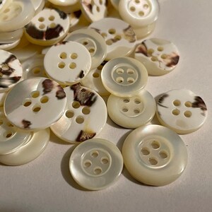 May include: A pile of white and brown speckled buttons with four holes each. The buttons are made of a smooth, shiny material.