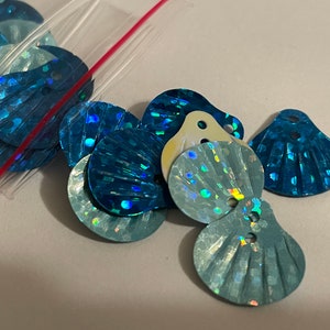 May include: A close-up of blue iridescent sequins shaped like seashells. The sequins have a holographic finish and are perfect for adding a touch of sparkle to any craft project.