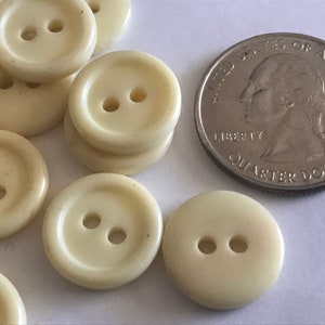 May include: A collection of off-white, round buttons with two holes each. A U.S. quarter coin is included for size comparison. The buttons are made of a smooth material and are slightly glossy.