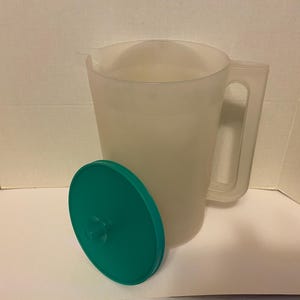 May include: A translucent, frosted plastic pitcher with a sturdy handle and a pouring spout. Beside the pitcher sits a round, solid teal-colored lid featuring a small, centered knob for easy handling. The pitcher has a cylindrical body shape.
