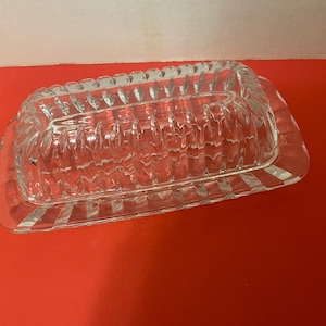 May include: A clear, rectangular glass butter dish with a ridged design. The dish has a lid and a base, both with a decorative, textured pattern. The dish is designed to hold butter and is suitable for kitchen use.