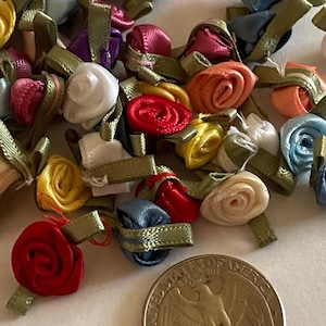 May include: A collection of small, colorful satin rosettes with green ribbon tails. The rosettes are arranged in a random pattern on a white surface.