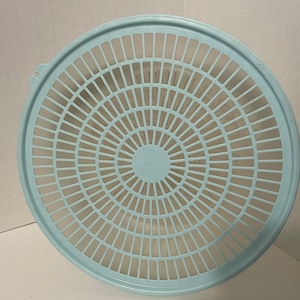 May include: A round, light blue plastic colander or strainer lid featuring a radial grid pattern of rectangular cutouts. The design includes a solid circular center and a raised outer rim with a small tab on the left side for easy handling.