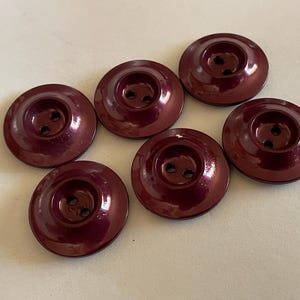May include: Seven burgundy-colored buttons with a glossy finish are arranged on a light surface. Each button is round with a recessed center and two small holes for sewing. The buttons are a deep, rich color.