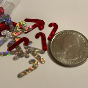 May include: A collection of red and silver candy cane-shaped confetti. The confetti has a holographic, iridescent finish. A U.S. quarter is included for size comparison. The confetti is likely for holiday or Christmas decorations.