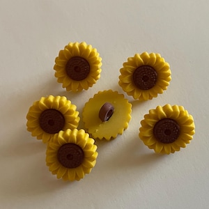 May include: A collection of six sunflower-shaped buttons. The buttons feature a yellow petal design with a brown center. One button is turned over, revealing a yellow back with a brown shank. These decorative buttons are ideal for sewing and craft projects.