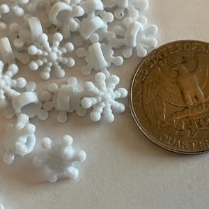 May include: A collection of small, white snowflake-shaped embellishments. A U.S. quarter coin is included for size comparison. The snowflakes have a three-dimensional design and are made of a smooth, possibly plastic, material. Ideal for craft projects.