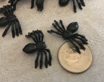 May include: A close-up of eight black plastic spiders. The spiders are about the size of a dime. They are scattered on a light gray surface.
