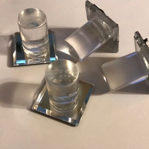 May include: Clear glass candle holders with mirrored bases. The candle holders are cylindrical and sit atop small, square mirrored bases. Two are upright, and two are tilted. The image is taken from above, with shadows cast on a white surface.
