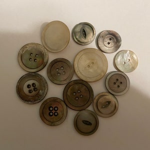May include: A collection of twelve round buttons in various shades of cream, gray, and brown. The buttons feature different designs, including some with four holes, some with two holes, and others with a single elongated center slot for attachment.