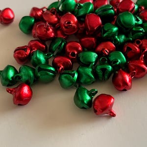 May include: A collection of small, shiny bells in red and green. The bells are scattered across a white surface. Each bell has a loop at the top and a slit on the side. The bells are likely for holiday decorations or crafts.