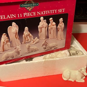May include: An 11-piece porcelain nativity set by Trimmings Etc. featuring white figurines with gold accents. The set includes figures of people and animals, displayed in a red box with the text PORCELAIN 11 PIECE NATIVITY SET printed on it.