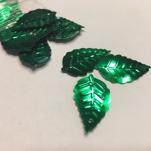 May include: Shiny, metallic green leaf-shaped embellishments scattered on a white surface. Some leaves are in a clear plastic bag. The leaves have detailed veining and a hole at the top for stringing or attaching.