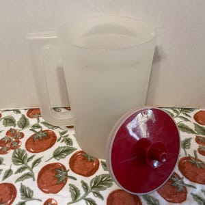 May include: A translucent plastic pitcher with a handle and a separate red lid. The pitcher is empty and sits on a tomato-patterned fabric. The lid is red with a central spout.