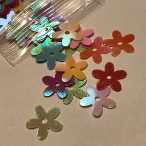 May include: Assortment of iridescent flower-shaped sequins in various colors, including pink, orange, yellow, green, and blue. The sequins are scattered on a white surface.