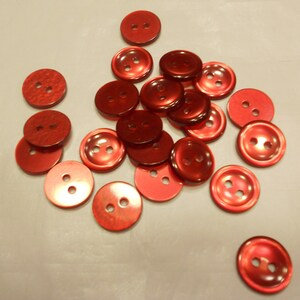 May include: A collection of shiny, red buttons with two or four holes each. The buttons are round and have a slightly iridescent finish, scattered on a light-colored surface. These buttons are suitable for sewing and craft projects.