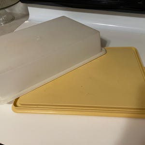 May include: A rectangular, translucent plastic container with a matching yellow lid. The lid is slightly ajar, revealing the interior. The container is designed for food storage and is placed on a white surface.