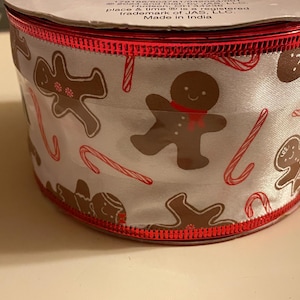 May include: A spool of white satin ribbon featuring a repeating pattern of brown gingerbread people and red and white candy canes. The ribbon has metallic red stitched edges and includes printed text on the top rim detailing manufacturing info.