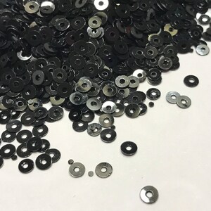 May include: A pile of black round sequins, scattered on a white surface. The sequins are shiny and have a metallic finish.