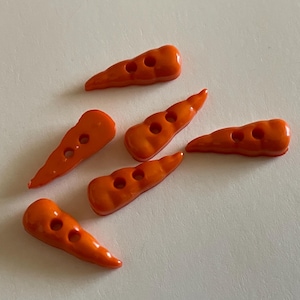 May include: Six orange carrot-shaped buttons with three holes each, scattered on a white surface. The buttons are glossy and have a slightly curved shape, resembling miniature carrots. Ideal for sewing and craft projects.