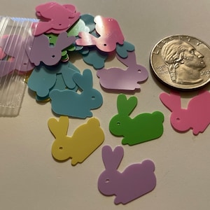 May include: A collection of colorful, plastic bunny-shaped confetti in pink, purple, blue, green, and yellow. A clear plastic bag contains more confetti. A US quarter coin is next to the confetti.