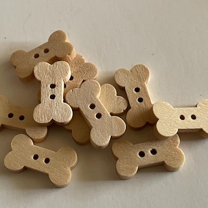 May include: A collection of light brown wooden buttons shaped like dog bones. Each button has two holes for sewing. The buttons are scattered on a light-colored surface.