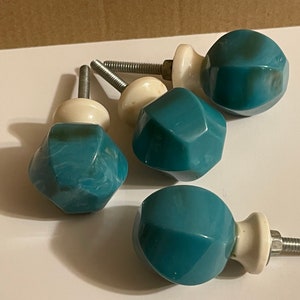 May include: A set of four cabinet knobs featuring faceted, teal-colored heads with white marbled swirls. Each knob has a smooth, rounded, off-white base and a silver-toned metal threaded bolt extending from the back for installation purposes.