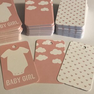 May include: Pink and white baby shower gift tags. Some tags feature a baby onesie graphic and the words "BABY GIRL". Other tags have cloud or heart patterns. The tags are rectangular with a hole at the top.