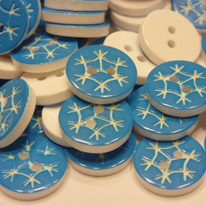May include: A collection of round buttons in white and bright blue. Some buttons are solid white with two holes, while others are blue with a white engraved snowflake pattern. The buttons are stacked and scattered, showing their circular shape and edges.