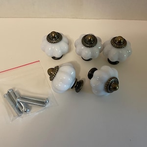 May include: Five white ceramic cabinet knobs with a flower-like design and bronze-colored accents. The knobs are accompanied by a small bag of silver screws, ready for installation. A decorative hardware item.