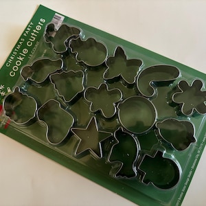 May include: A set of 15 Christmas-themed cookie cutters in various shapes, including stars, snowflakes, and ornaments. The cutters are made of shiny metal and are packaged on a green plastic tray. The packaging reads "Christmas Party Cookie Cutters".